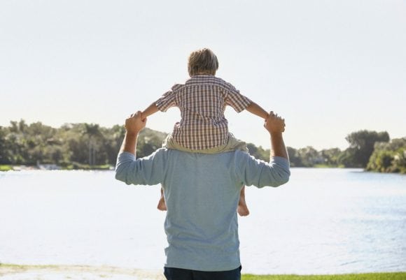 Father giving young son shoulder ride outdoors at park by a lake